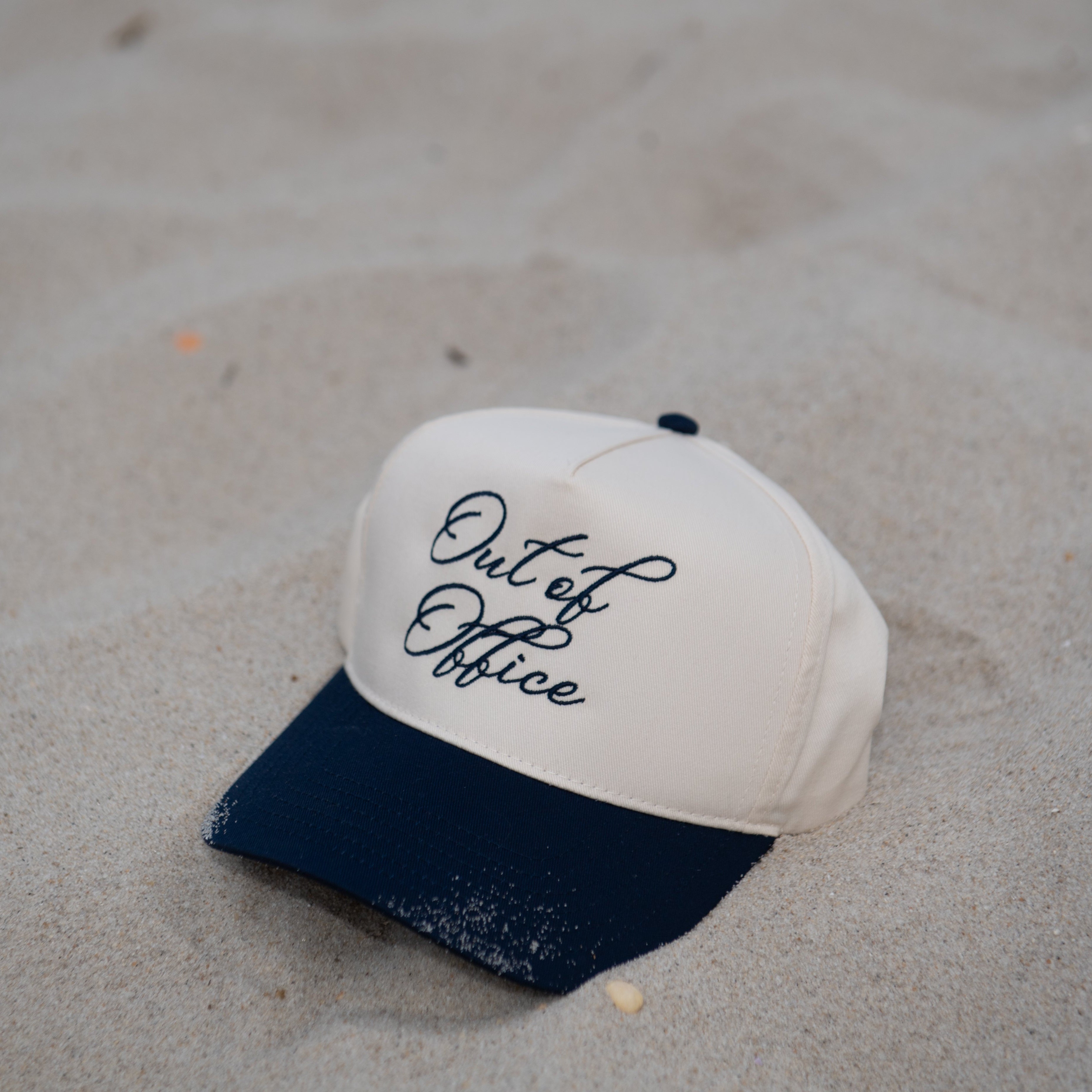 White cap with navy brim and 'Out of Office' text on sandy surface